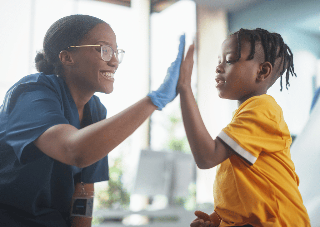 Young African American Boy Sitting In The Chair In Bright Hospital And Getting His Flu Vaccine. Female Black Nurse Is Finished Performing Injection. Professional Woman High-Fives A Kid For Being Brave