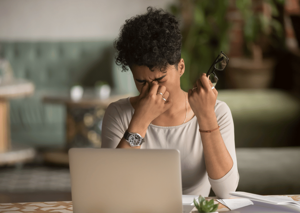 Overworked Black female at work, holding glasses, feeling eye strain, at a computer