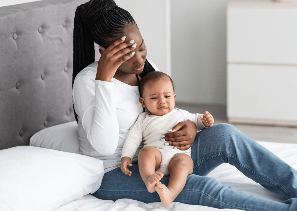 Postpartum Black woman sitting with her baby in a bed at home, feeling headache, touching forehead with hand. 